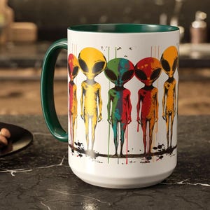 Puede incluir: Taza de cer&aacute;mica blanca con borde verde y un gr&aacute;fico colorido de cinco extraterrestres con pintura goteando. Los extraterrestres son amarillos, verdes, rojos, azules y naranjas.