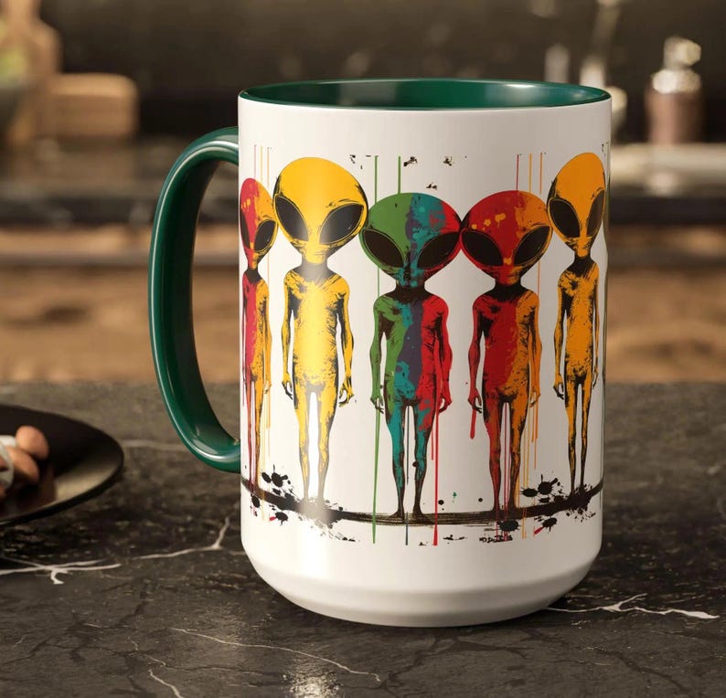 Puede incluir: Taza de cer&aacute;mica blanca con borde verde y un gr&aacute;fico colorido de cinco extraterrestres con pintura goteando. Los extraterrestres son amarillos, verdes, rojos, azules y naranjas.