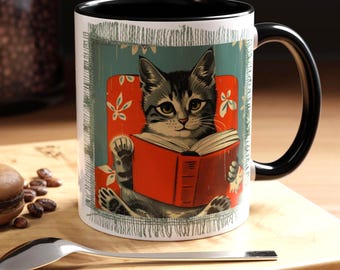 May include: A white ceramic mug with a black rim and handle features a vintage illustration of a cat sitting in a chair and reading a red book. The cat has gray and white fur and is looking directly at the viewer.