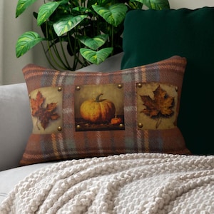 Op de afbeelding: Rechthoekig decoratief kussen met een ruitpatroon in bruin, oranje en groen. Het kussen heeft drie rechthoekige panelen met afbeeldingen van herfstbladeren en pompoenen. De panelen zijn bevestigd met koperkleurige klinknagels.