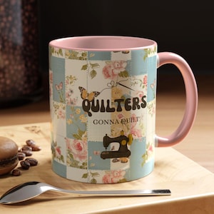 Op de afbeelding: Een keramische mok met een roze handvat en binnenkant. De buitenkant heeft een patchwork-ontwerp met bloempatronen en vlinders. De tekst op de mok luidt "Quilters Gonna Quilt" met een illustratie van een naaimachine.