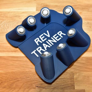 Op de afbeelding: Een blauwe Rev Trainer met zeven cilindrische houders voor batterijen. De tekst "REV TRAINER" is in het wit op het blauwe oppervlak gedrukt. Het apparaat is ontworpen om batterijen te bevatten.
