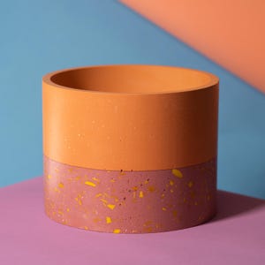Può includere: Un vaso cilindrico con un design a due toni. La metà superiore è di colore arancione pieno, mentre la metà inferiore è di colore rosa con macchie dorate.