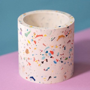 Op de afbeelding: Een witte cilindrische plantenpot met een gespikkeld terrazzo-patroon in verschillende kleuren, waaronder roze, blauw, groen, geel en oranje.