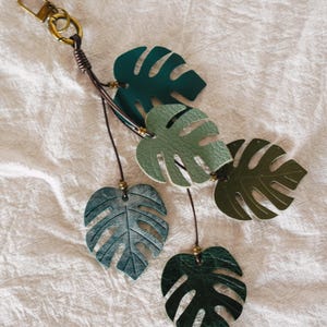 Puede incluir: Un llavero con cinco dijes de cuero en forma de hoja de monstera en varios tonos de verde, incluyendo verde bosque oscuro, salvia y oliva. Las hojas est&aacute;n unidas a finos cordones de cuero marr&oacute;n con peque&ntilde;as cuentas de lat&oacute;n y un mosquet&oacute;n.