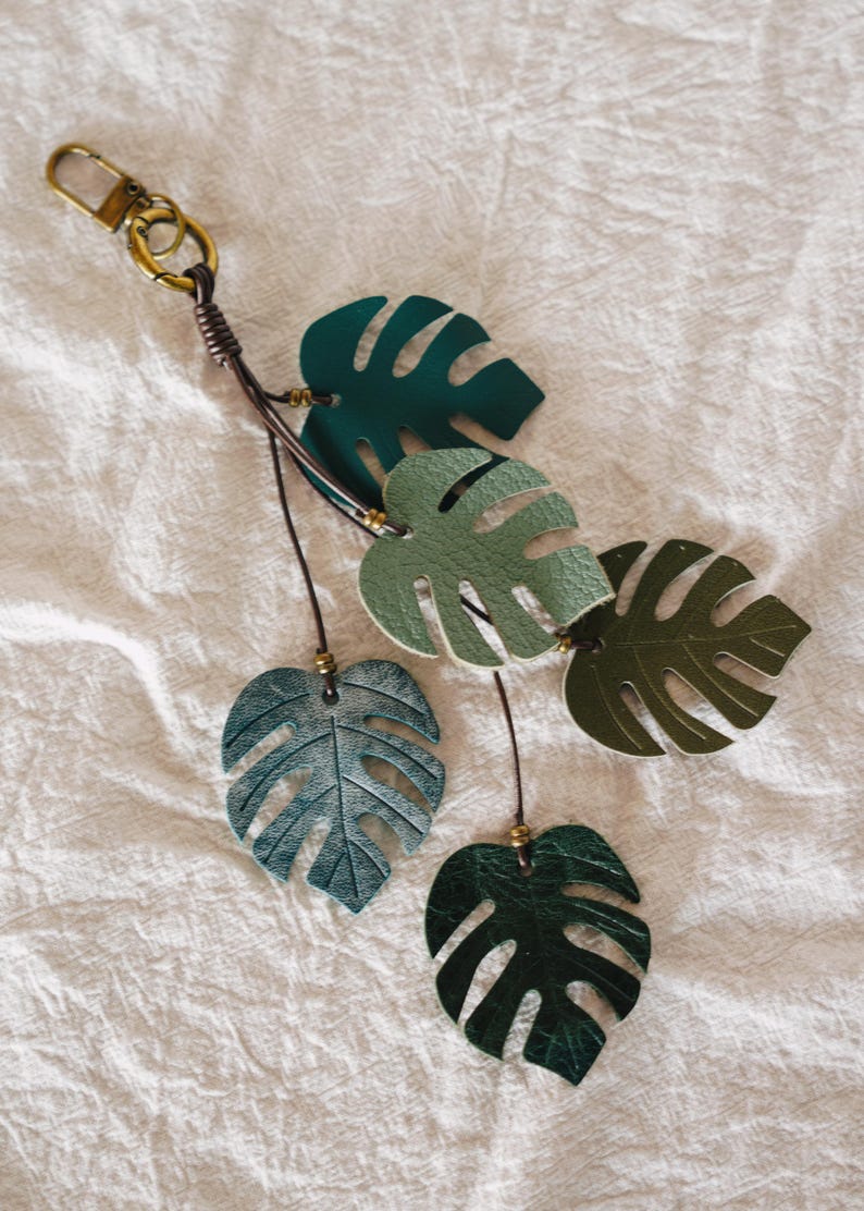 Puede incluir: Un llavero con cinco dijes de cuero en forma de hoja de monstera en varios tonos de verde, incluyendo verde bosque oscuro, salvia y oliva. Las hojas est&aacute;n unidas a finos cordones de cuero marr&oacute;n con peque&ntilde;as cuentas de lat&oacute;n y un mosquet&oacute;n.