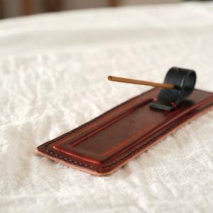 May include: A brown leather incense holder with a black incense stick holder. The rectangular tray has a stitched border and a raised center. An incense stick is placed in the holder. The background is a white, textured surface.