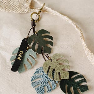 Puede incluir: Un llavero decorativo con múltiples recortes de hojas de monstera de cuero en tonos verde oscuro, salvia y azul grisáceo. Una etiqueta de cuero negro con el texto dorado "Odd" está unida a un anillo de metal color bronce y un cordón marrón.