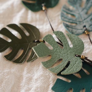 Puede incluir: Una colecci&oacute;n de recortes con forma de hoja de monstera hechos de varios tonos de cuero verde. Cada hoja presenta recortes distintivos y est&aacute; unida a un cord&oacute;n fino de color marr&oacute;n oscuro con peque&ntilde;as cuentas met&aacute;licas decorativas.