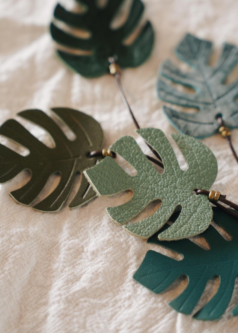 Puede incluir: Una colecci&oacute;n de recortes con forma de hoja de monstera hechos de varios tonos de cuero verde. Cada hoja presenta recortes distintivos y est&aacute; unida a un cord&oacute;n fino de color marr&oacute;n oscuro con peque&ntilde;as cuentas met&aacute;licas decorativas.