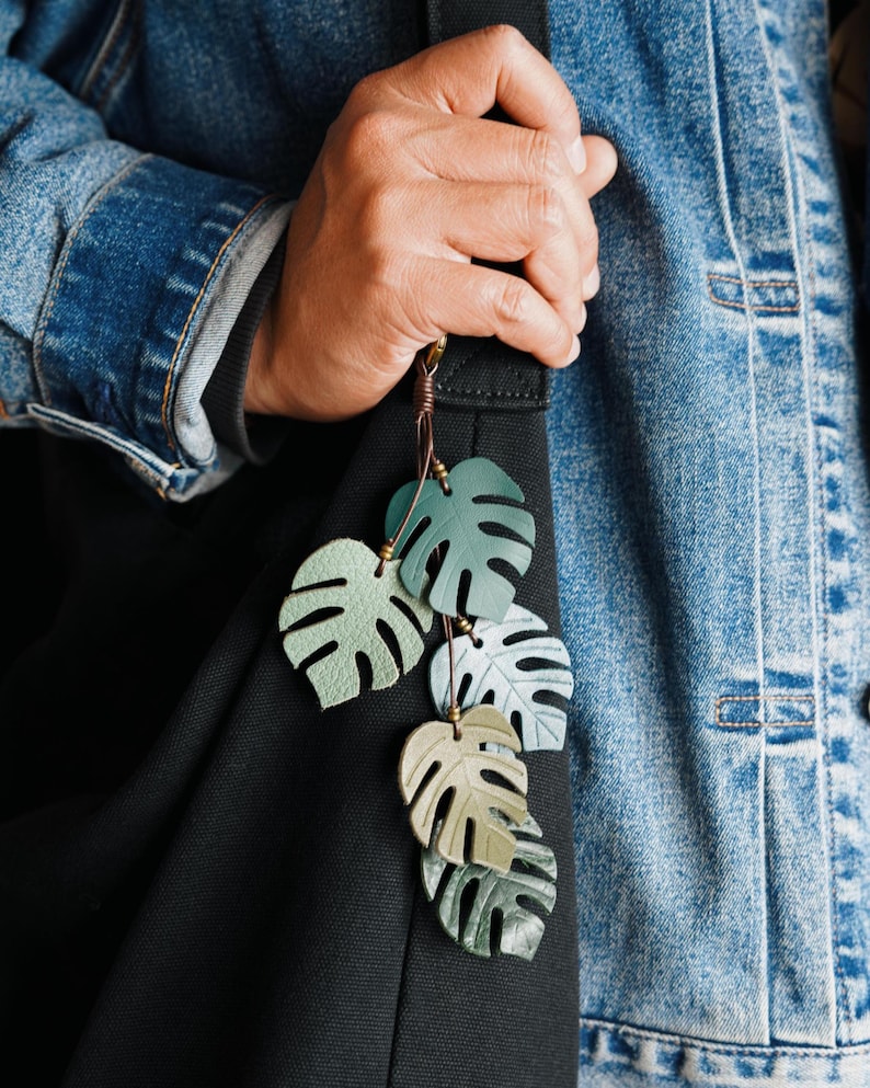 Puede incluir: Un colgante para bolso con cinco recortes de hojas de monstera superpuestos en varios tonos de verde, desde salvia claro hasta verde bosque oscuro. Las hojas est&aacute;n unidas a un cord&oacute;n de cuero marr&oacute;n oscuro con peque&ntilde;as cuentas met&aacute;licas.