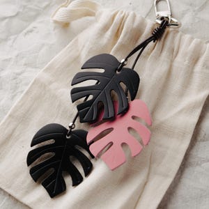 Puede incluir: Un llavero con tres dijes en forma de hoja de monstera unidos a un cierre de metal plateado. Dos de las hojas son de color negro mate y una hoja es de color rosa claro. Los dijes cuelgan de finos cordones de color marrón oscuro con cuentas.