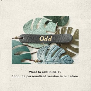 Puede incluir: Una colecci&oacute;n de recortes de cuero con forma de hoja de monstera en varios tonos de verde, incluyendo menta, bosque y oliva. Una tira de cuero gris oscuro se superpone, con el texto "Odd" grabado en oro y peque&ntilde;as cuentas met&aacute;licas.