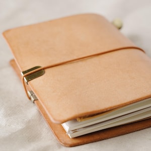 May include: A tan leather notebook cover featuring a smooth, natural finish. The cover is secured by a thin, elastic cord that wraps horizontally around the exterior, held in place by a small, metallic brass-colored clip on the left side.