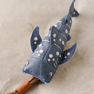 Puede incluir: Una figura de tiburón ballena azul hecha de cuero texturizado con patrones de lunares blancos. El tiburón presenta costuras azules visibles a lo largo de los bordes, dos pequeños remaches metálicos plateados como ojos y un cordón de cuero marrón.
