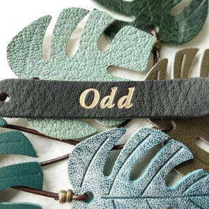Puede incluir: Una etiqueta de cuero negro con la palabra Odd grabada en oro, superpuesta sobre varias formas de hojas de cuero texturizado en varios tonos de verde, incluyendo menta, bosque y oliva, conectadas por cordones de cuero marr&oacute;n y cuentas de lat&oacute;n.