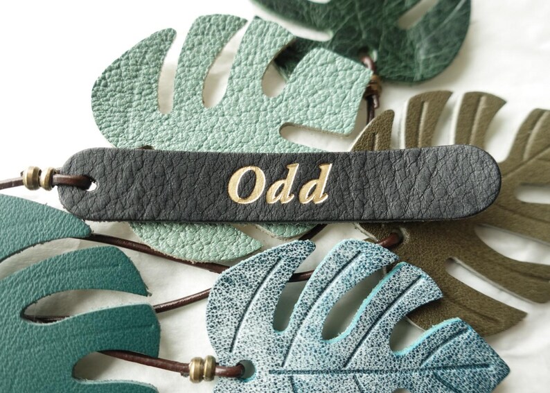 Puede incluir: Una etiqueta de cuero negro con la palabra Odd grabada en oro, superpuesta sobre varias formas de hojas de cuero texturizado en varios tonos de verde, incluyendo menta, bosque y oliva, conectadas por cordones de cuero marr&oacute;n y cuentas de lat&oacute;n.