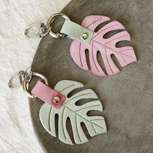 May include: Two keychains with monstera leaf designs. One leaf is light pink, the other is light green. Each leaf is attached to a silver-colored metal ring and clasp with a matching leather strap. The keychains are on a gray surface.