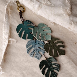May include: A keychain featuring five die-cut monstera leaf shapes in varying shades of green and blue-green. The leaves are attached to a dark brown cord with small metallic beads and connected to a brass-toned metal lobster clasp and ring.