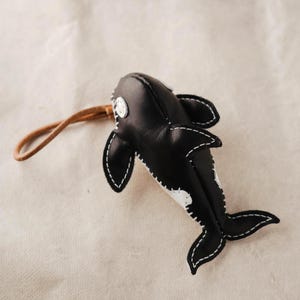 May include: A black and white leather orca whale keychain featuring visible white stitching along the edges, a smooth finish, and a tan leather cord loop attached to the top for hanging or attachment to personal items.
