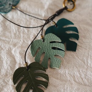 Puede incluir: Un llavero con varios recortes en forma de hoja de monstera en diferentes tonos de verde y azul. Las hojas est&aacute;n unidas a un cord&oacute;n marr&oacute;n oscuro con peque&ntilde;as cuentas met&aacute;licas y un mosquet&oacute;n y anilla de metal en tono lat&oacute;n.