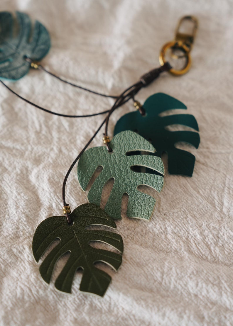 Puede incluir: Un llavero con varios recortes en forma de hoja de monstera en diferentes tonos de verde y azul. Las hojas est&aacute;n unidas a un cord&oacute;n marr&oacute;n oscuro con peque&ntilde;as cuentas met&aacute;licas y un mosquet&oacute;n y anilla de metal en tono lat&oacute;n.