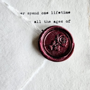 May include: A burgundy wax seal with a rose design, resting on a textured white paper with the handwritten text "Daniel," and a portion of a typed message. The seal is circular and features a detailed floral motif.
