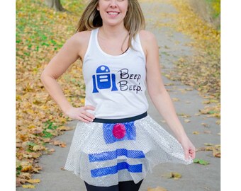Puede incluir: Una mujer lleva una camiseta sin mangas blanca con un gráfico de R2-D2 azul y blanco y el texto "Beep Beep". También lleva una falda de lentejuelas plateadas con rayas azules y blancas y un círculo rojo. La falda está diseñada para parecerse a R2-D2.
