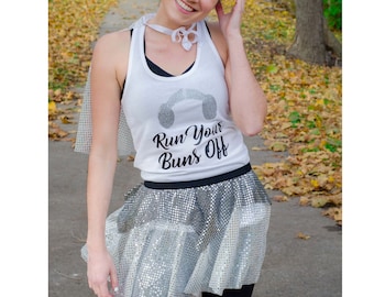 Puede incluir: Un top blanco sin mangas con el texto "Run Your Buns Off" y un gráfico de auriculares. La modelo lleva una falda de lentejuelas plateadas y tiene el pelo en dos moños.