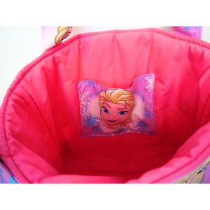 May include: A bright pink fabric tote bag with a quilted interior lining. The center of the bag features a rectangular patch displaying a graphic illustration of a blonde character with blue eyes, surrounded by blue snowflake patterns on a pink background.