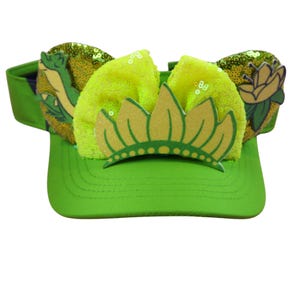 May include: A green visor with a yellow bow and a gold crown design. The visor has sequined ears with a cartoon alligator and a flower. The visor is a bright green colour.