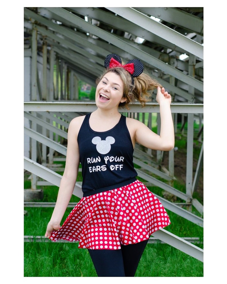 May include: A woman wearing a black tank top with the text "Run Your Ears Off" and a white Mickey Mouse silhouette. She is also wearing a red and white polka dot skirt and a black headband with red bow and black Mickey Mouse ears.