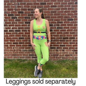 Puede incluir: Una mujer lleva un top verde sin mangas con un cintur&oacute;n dorado y un dise&ntilde;o estampado verde y blanco. Tambi&eacute;n lleva leggings verdes con una cintura colorida. Los leggings son cortos y tienen un peque&ntilde;o desgarro en la pierna izquierda. El texto "Leggings vendidos por separado" est&aacute; en la parte inferior de la imagen.