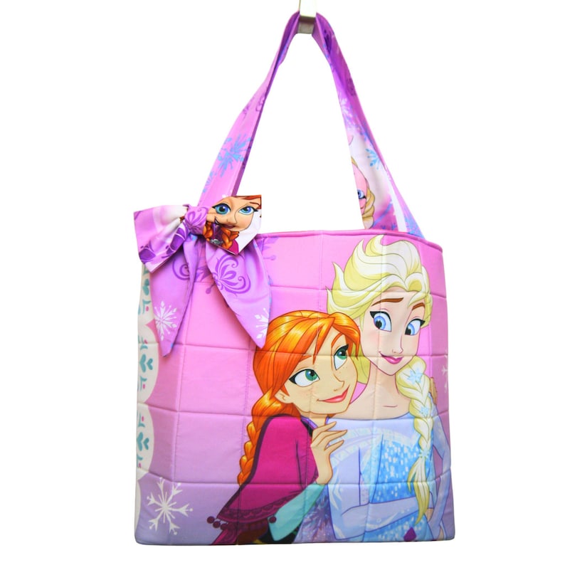 May include: A quilted tote bag in shades of pink and purple featuring a graphic print of two animated characters with red and blonde hair. The bag includes a large decorative bow on the side and two shoulder straps with a snowflake pattern.