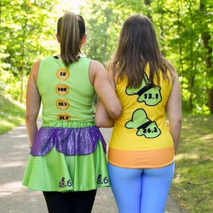 Puede incluir: Dos personas que llevan camisetas de correr verdes y amarillas con n&uacute;meros. Las camisetas est&aacute;n inspiradas en un personaje de dibujos animados. La persona de la izquierda lleva una falda verde con lentejuelas moradas. Est&aacute;n caminando por un sendero en un bosque.