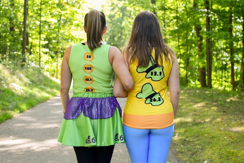 Puede incluir: Dos personas que llevan camisetas de correr verdes y amarillas con n&uacute;meros. Las camisetas est&aacute;n inspiradas en un personaje de dibujos animados. La persona de la izquierda lleva una falda verde con lentejuelas moradas. Est&aacute;n caminando por un sendero en un bosque.