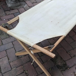 May include: A white canvas cot with wooden frame. The cot is folded and sitting on a brick patio.