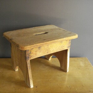 May include: A small, rectangular wooden stool with a natural light brown finish. The top surface features a centered, elongated oval cutout handle. The stool is supported by four sturdy legs with arched cutouts between them on the sides.