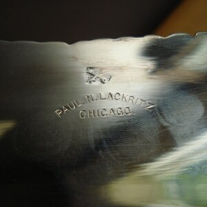 May include: Close-up of a silver metal object with a scalloped edge. The text "PAUL N. LACKRITZ CHICAGO" is engraved on the surface. A small emblem is also visible above the text.