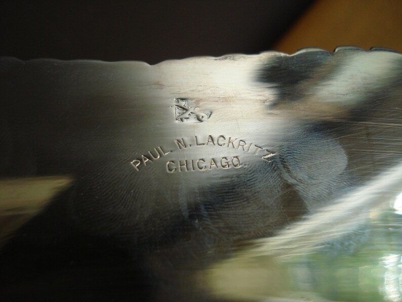 May include: Close-up of a silver metal object with a scalloped edge. The text "PAUL N. LACKRITZ CHICAGO" is engraved on the surface. A small emblem is also visible above the text.