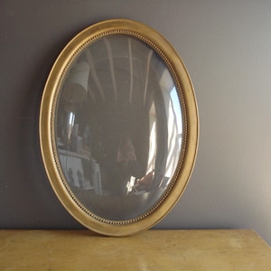 May include: An oval-shaped, antique gold picture frame with a clear glass front. The frame has a beaded edge and a slightly domed glass. The frame is resting on a light-colored surface against a dark gray wall.