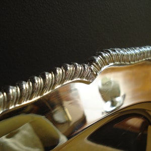 May include: Close-up of a silver-colored serving dish with a decorative, ridged edge. The dish has a reflective surface, and the edge has a wavy design. The background is dark, highlighting the bright metal.