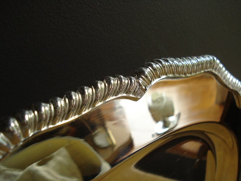 May include: Close-up of a silver-colored serving dish with a decorative, ridged edge. The dish has a reflective surface, and the edge has a wavy design. The background is dark, highlighting the bright metal.