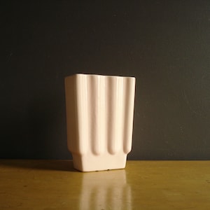 Peut inclure: Un vase en céramique rose pâle de forme rectangulaire avec des détails verticaux côtelés. Le vase a une base légèrement évasée et une finition lisse et mate. C'est un objet décoratif.