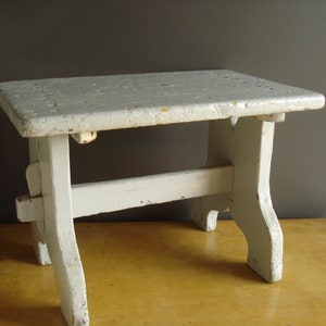 May include: A small, white wooden stool with a simple design. The stool has a rectangular top and four legs that are slightly curved at the bottom.