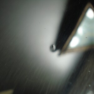 May include: Abstract macro shot featuring a reflective, dark gray surface with subtle scratches. A blurred, triangular shape with bright highlights is visible in the background. A small water droplet is in the foreground.