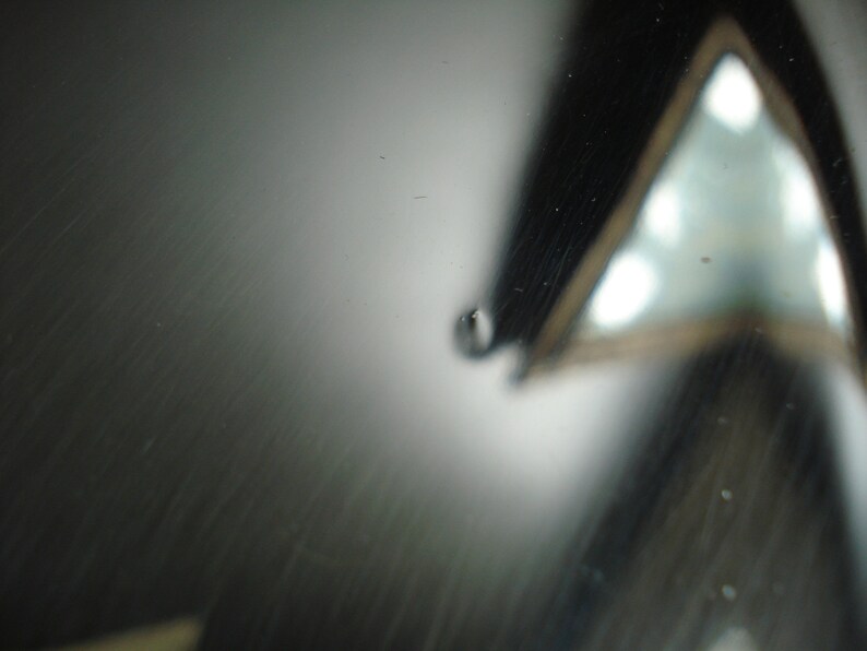 May include: Abstract macro shot featuring a reflective, dark gray surface with subtle scratches. A blurred, triangular shape with bright highlights is visible in the background. A small water droplet is in the foreground.