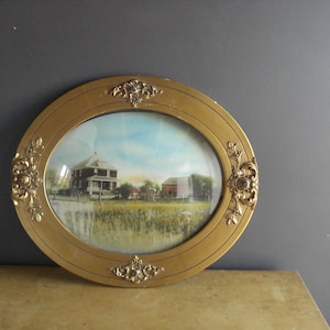 May include: An oval, antique gold-colored picture frame with an image of a house and barn in a field. The frame has ornate floral detailing at the corners and along the sides. The image inside the frame depicts a rural landscape with a house and barn.