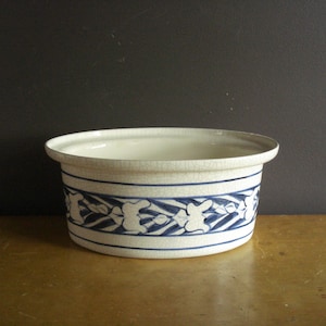 May include: A round ceramic bowl with a cream-colored base and a fine crackle glaze finish. The exterior features a horizontal band decorated with a repeating blue painted pattern of stylized leaves and geometric shapes around the center.
