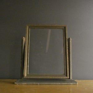 May include: An antique, rectangular picture frame with a gold-colored border and a clear glass pane. The frame is supported by two textured, vertical posts and a rectangular base. The frame is likely made of wood and sits on a wooden surface.